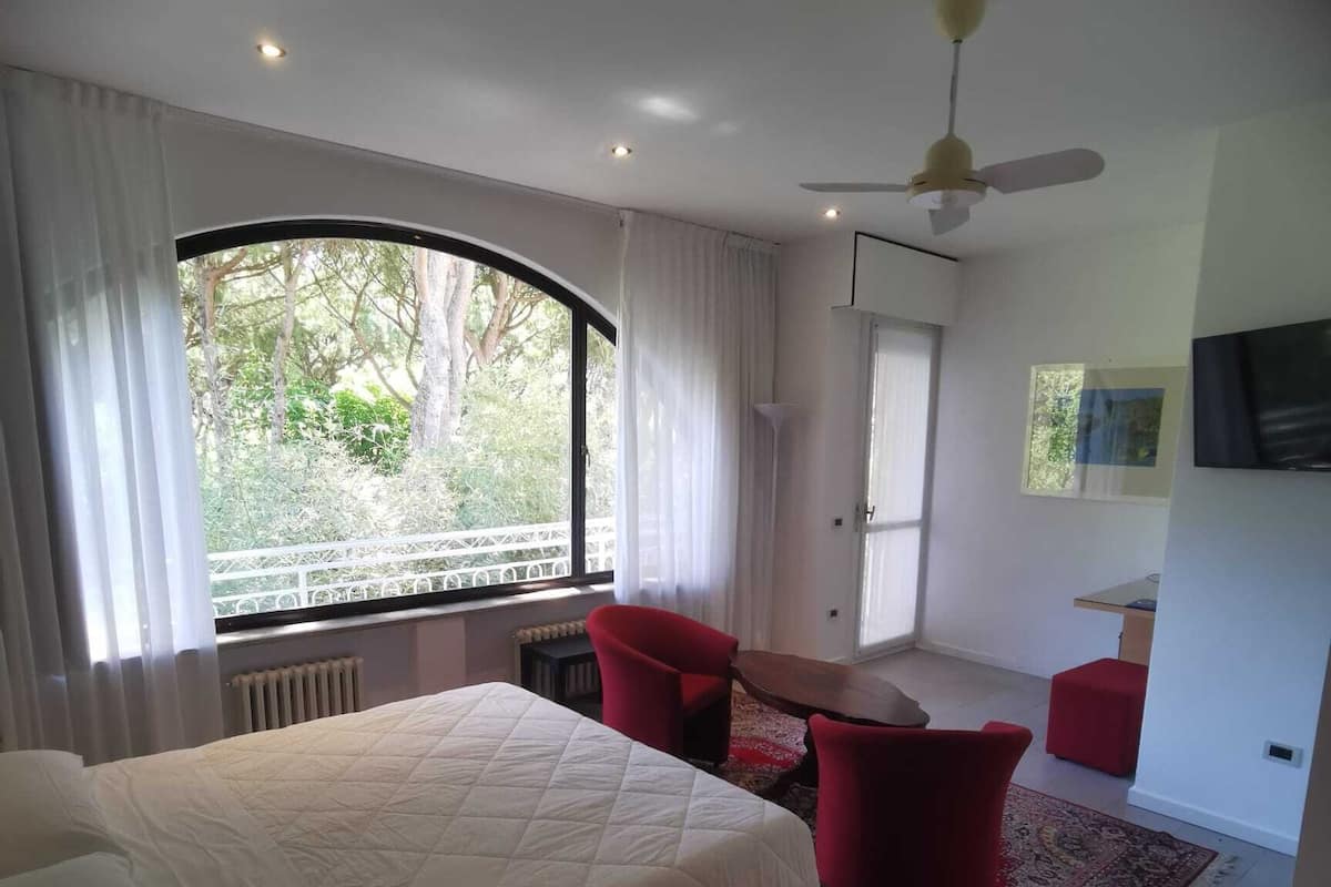 A hotel room with a large bed, a red chair, a small table, and a view of greenery outside.