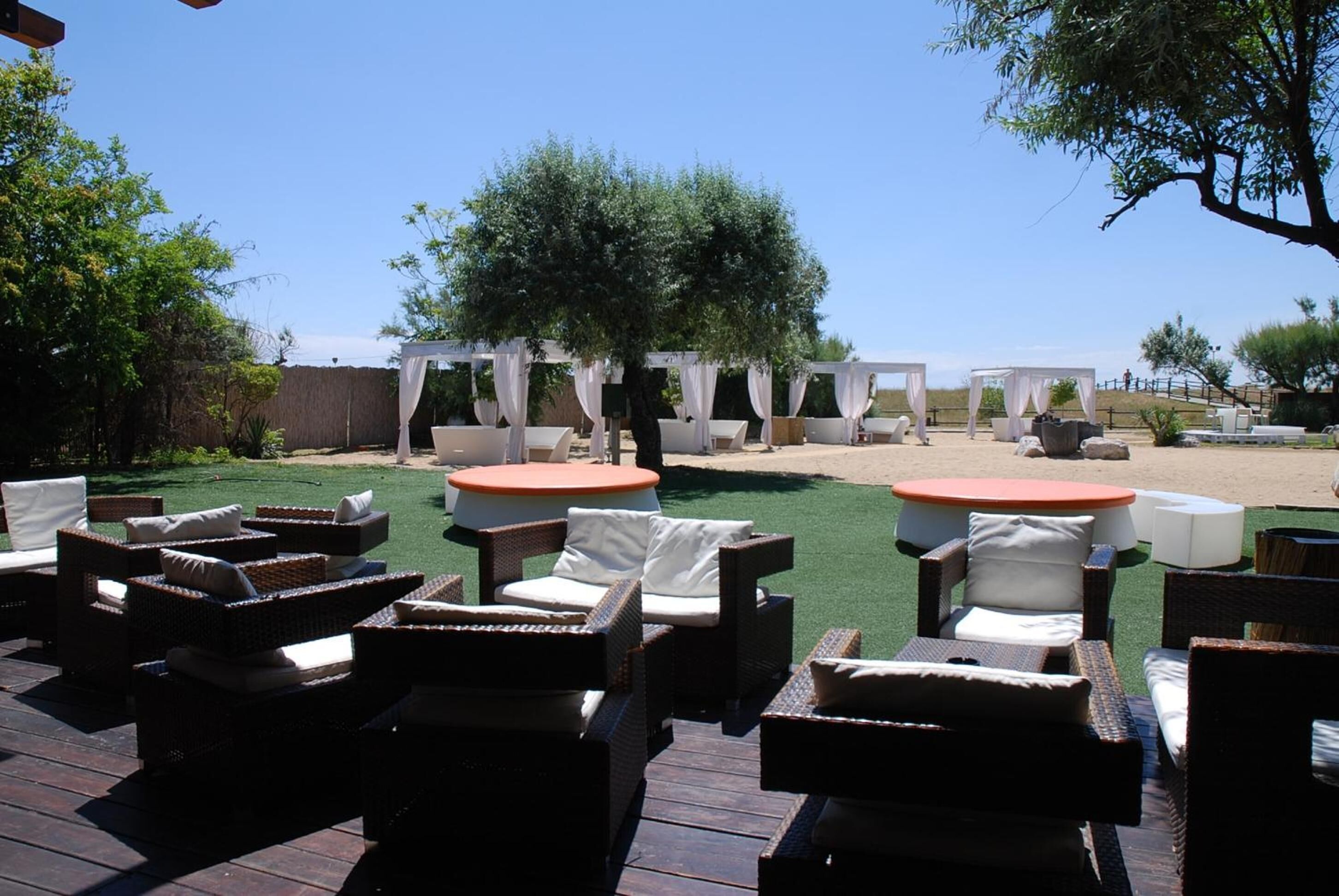 Ein möblierter Terrassenbereich mit Korbmöbeln und weißen Kissen, einem runden Tisch mit orangefarbener Tischplatte und einer Pergola mit weißen Vorhängen.