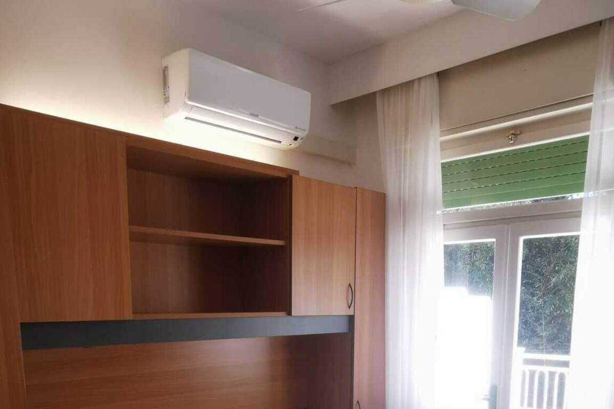 A bedroom with a bed, wooden headboard, built-in shelves, a window with curtains, and an air conditioning unit.
