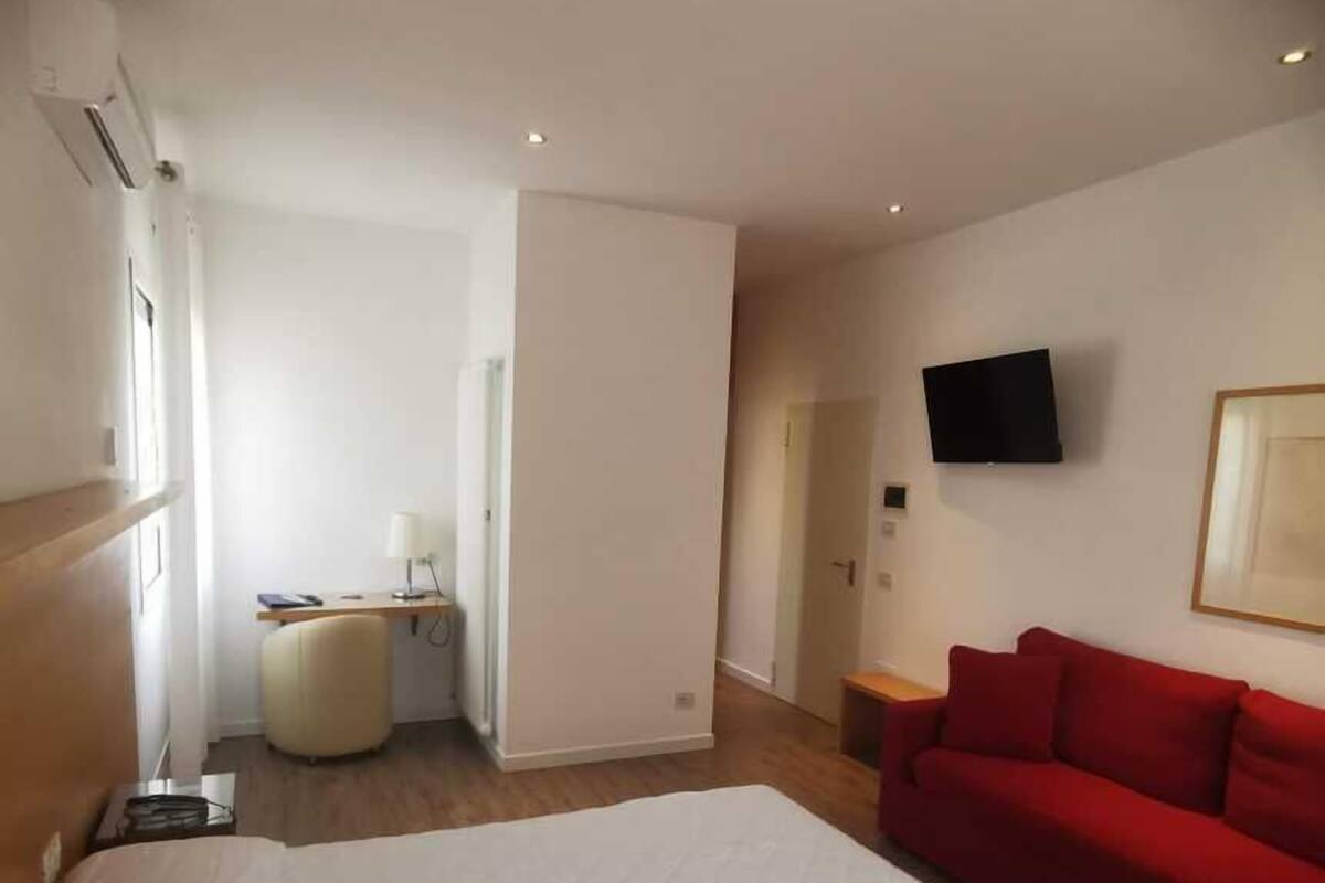 A hotel room with a large bed, a red sofa, a TV mounted on the wall, and a desk with a lamp.