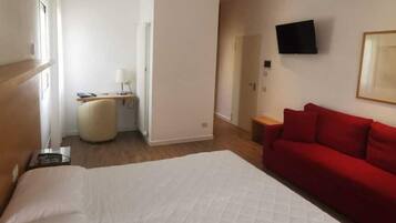 A hotel room with a large bed, a red sofa, a TV mounted on the wall, and a desk with a lamp.