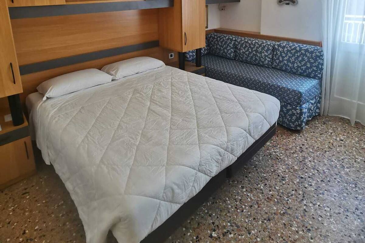A bedroom with a bed, a sofa, wooden cabinets, an air conditioning unit, and a small decorative mirror.