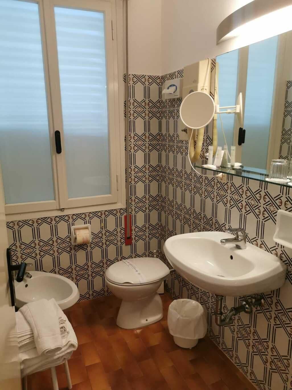 Et badeværelse med toilet, vask og spejl. Der er et vindue med skodder, tapet med mønster og en håndklædestang.