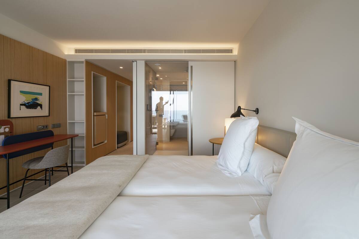 Ein modernes Hotelzimmer mit einem Bett, einem Schreibtisch und einem Sessel. Eine Tür führt zu einem Badezimmer und einem Flur.