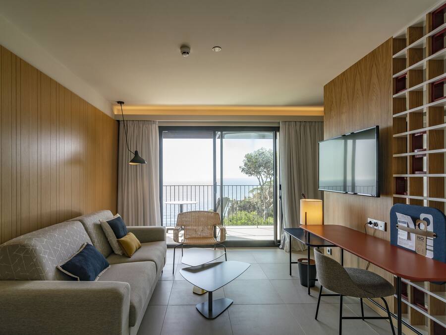 Una habitación de hotel moderna con sofá, mesa de comedor, una silla, televisión y balcón con vistas.