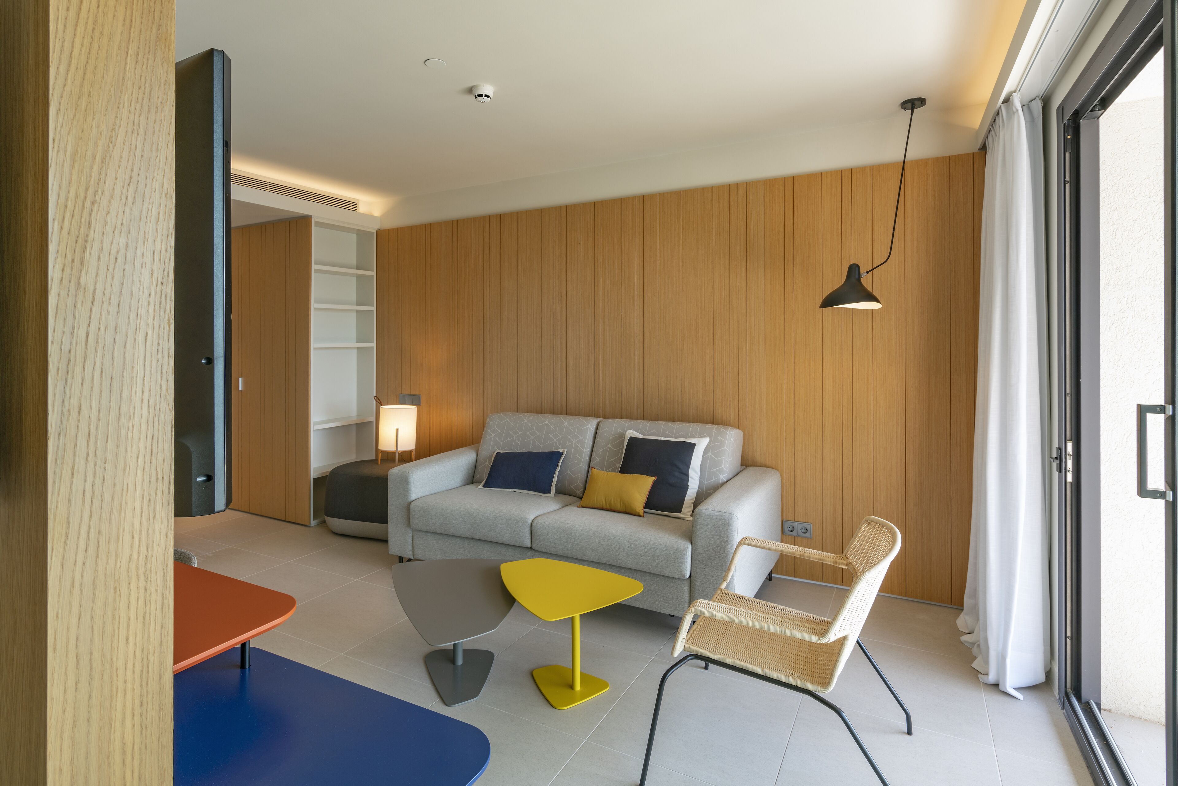 Een moderne woonkamer met een grijze bank, een gele bijzettafel, een rieten stoel en een zwarte hanglamp.
