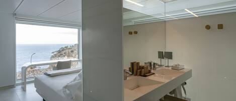 A modern bedroom with a large bed, a bathroom with a large sink and mirrors, and a view of the sea.