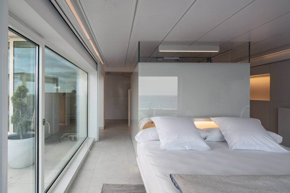 Ein modernes Schlafzimmer mit einem großen Bett, einer Glastür, die auf einen Balkon führt, und Blick ins Freie.