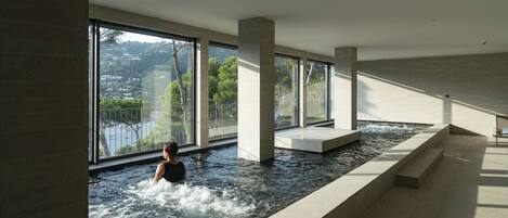 Ein modernes Hallenbad mit großen Fenstern, Blick auf eine Hügelkette und einer ruhigen Wasseroberfläche.
