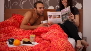 Una pareja en una cama, con una revista, una taza y un plato con comida y bebida.