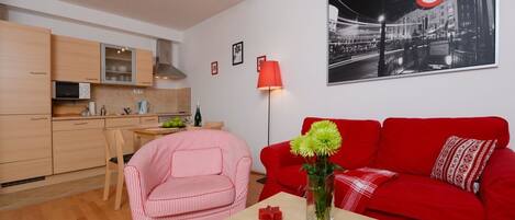 A compact living space with a kitchenette, a red sofa, a striped chair, and a table with a vase and remote controls.