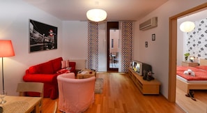 Un salón moderno con un sofá rojo, una mesita y un sillón rojo. El suelo es de madera, hay un televisor montado en la pared y una puerta que da a otra habitación.