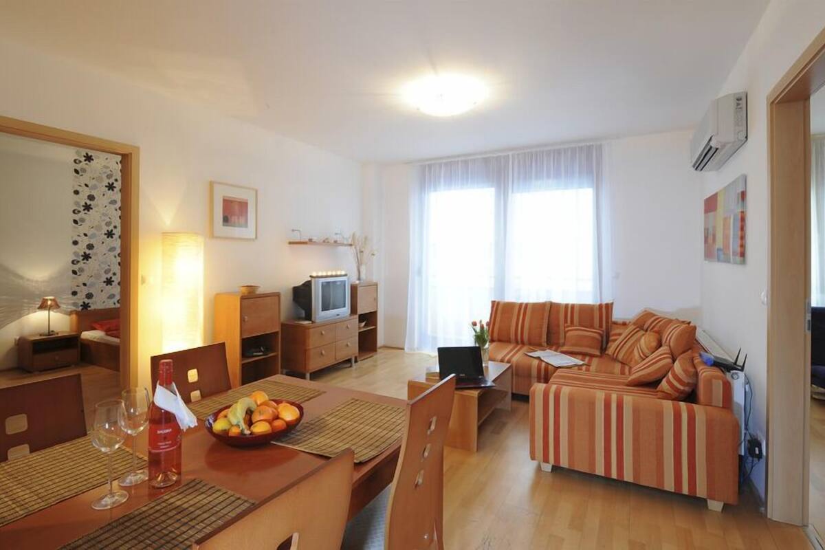 Ein helles Wohnzimmer mit einer Couch, einem Esstisch mit Stühlen, einem Fernseher und einer Lampe.