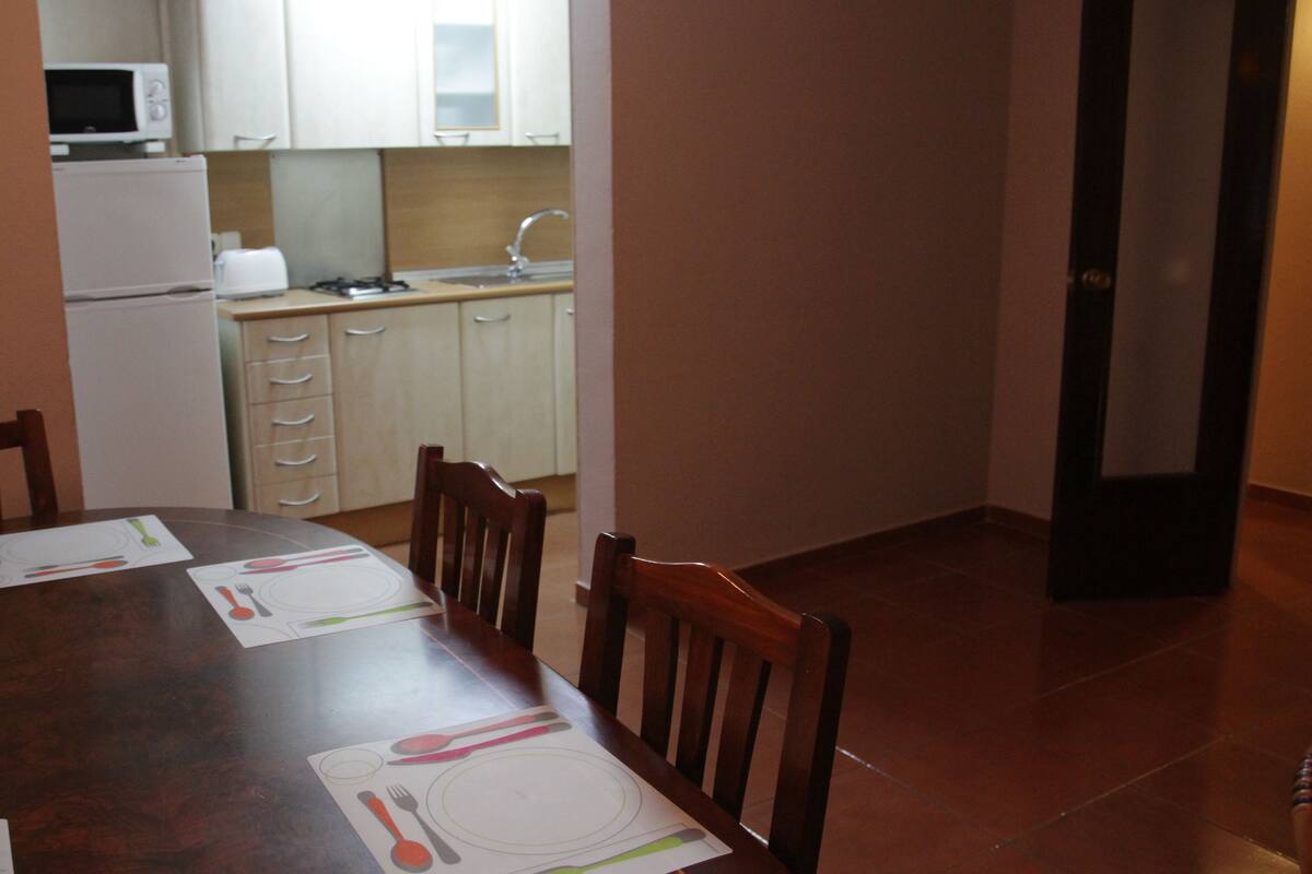 Un comedor con una mesa de madera dispuesta para cuatro, una cocina al fondo con armarios blancos y un microondas, y un piso de baldosas.