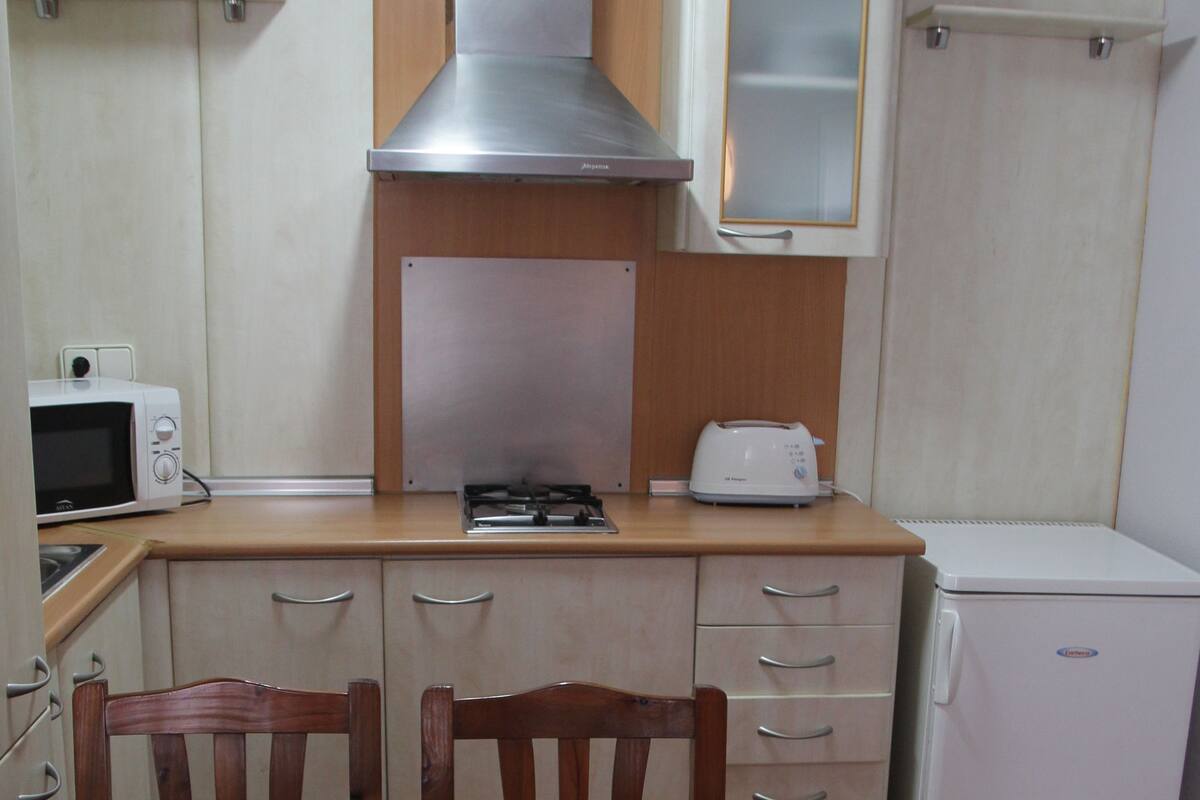Cocina compacta con estufa, microondas, tostadora y refrigerador. Hay sillas de madera alrededor de una mesa con un mantel individual.