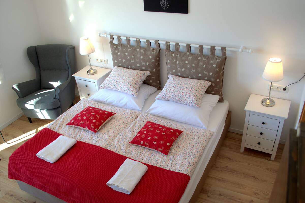 A bed with a red blanket and pillows, two bedside tables with lamps, a grey armchair, and a white headboard.