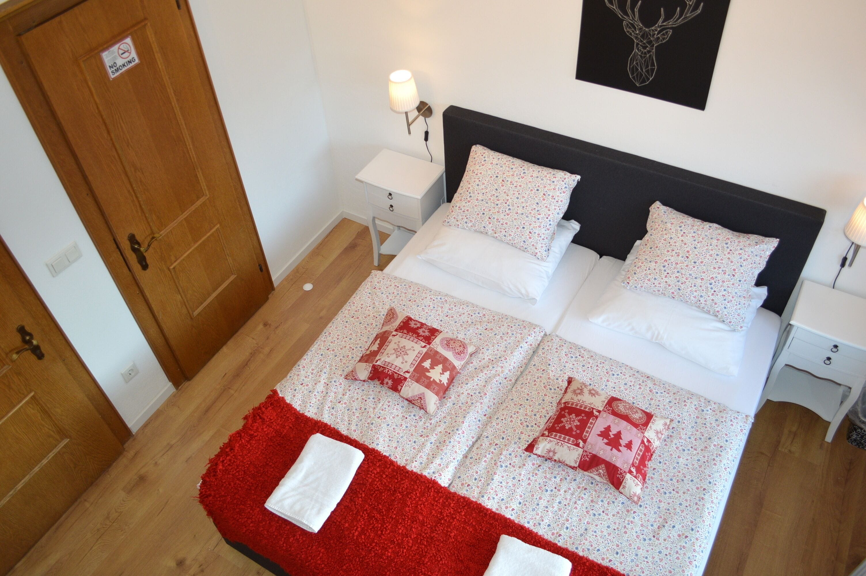 A neatly made bed with a red and white blanket, two bedside tables with lamps, a wooden door, and a wall-mounted deer head decoration.