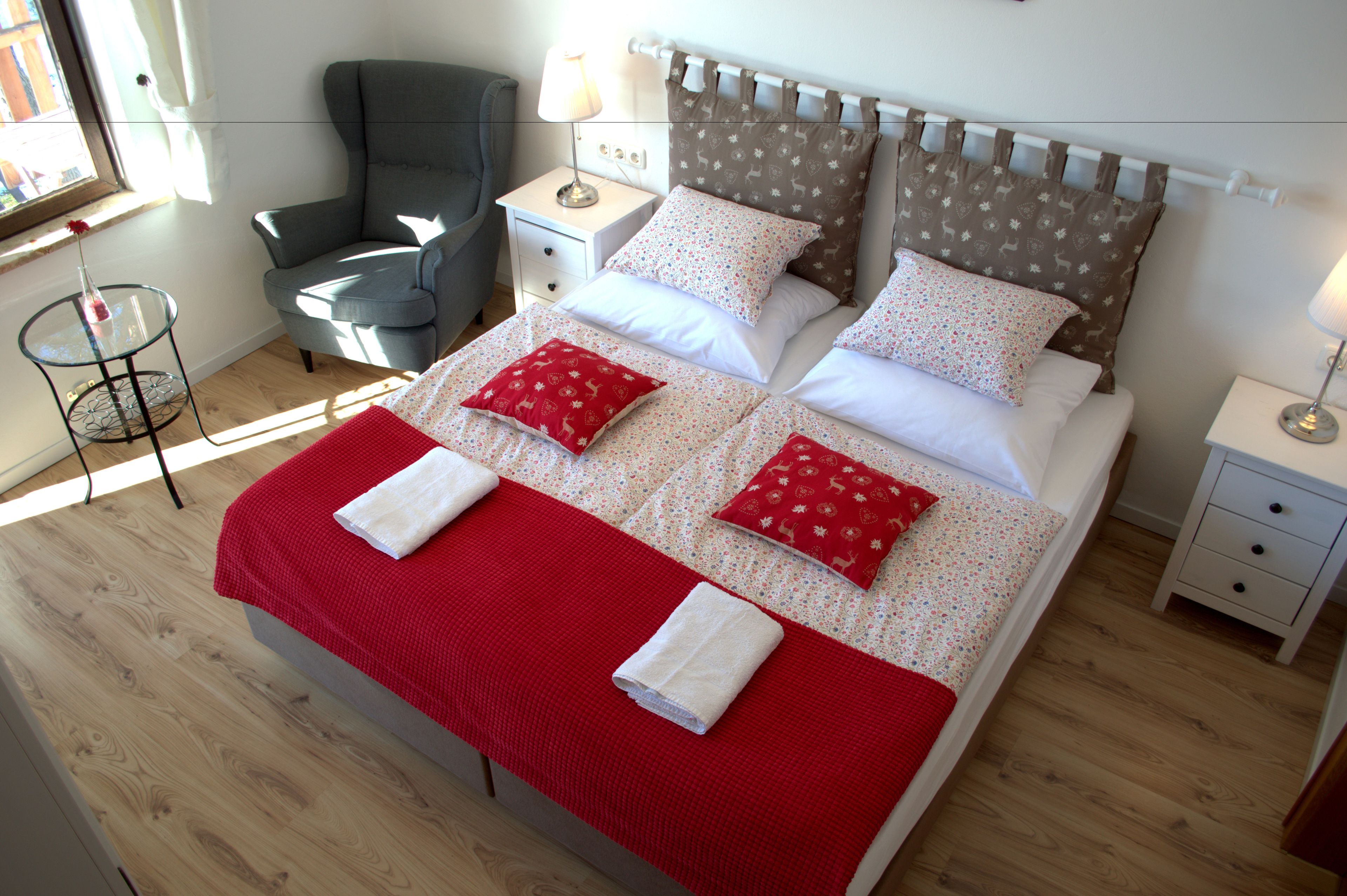 A neatly made bed with a red blanket and pillows, a grey armchair, and a small table with a lamp.