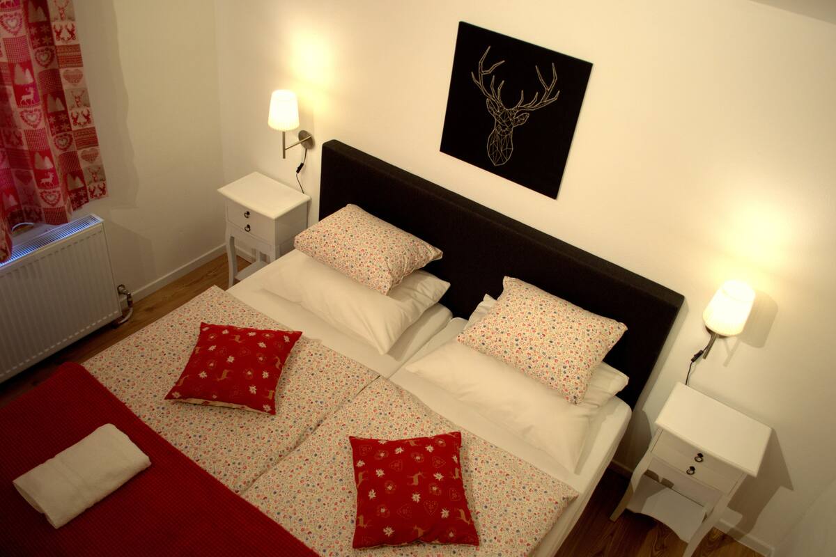 A neatly made bed with floral bedding and red pillows, a headboard with a deer head artwork, bedside lamps, and a radiator.