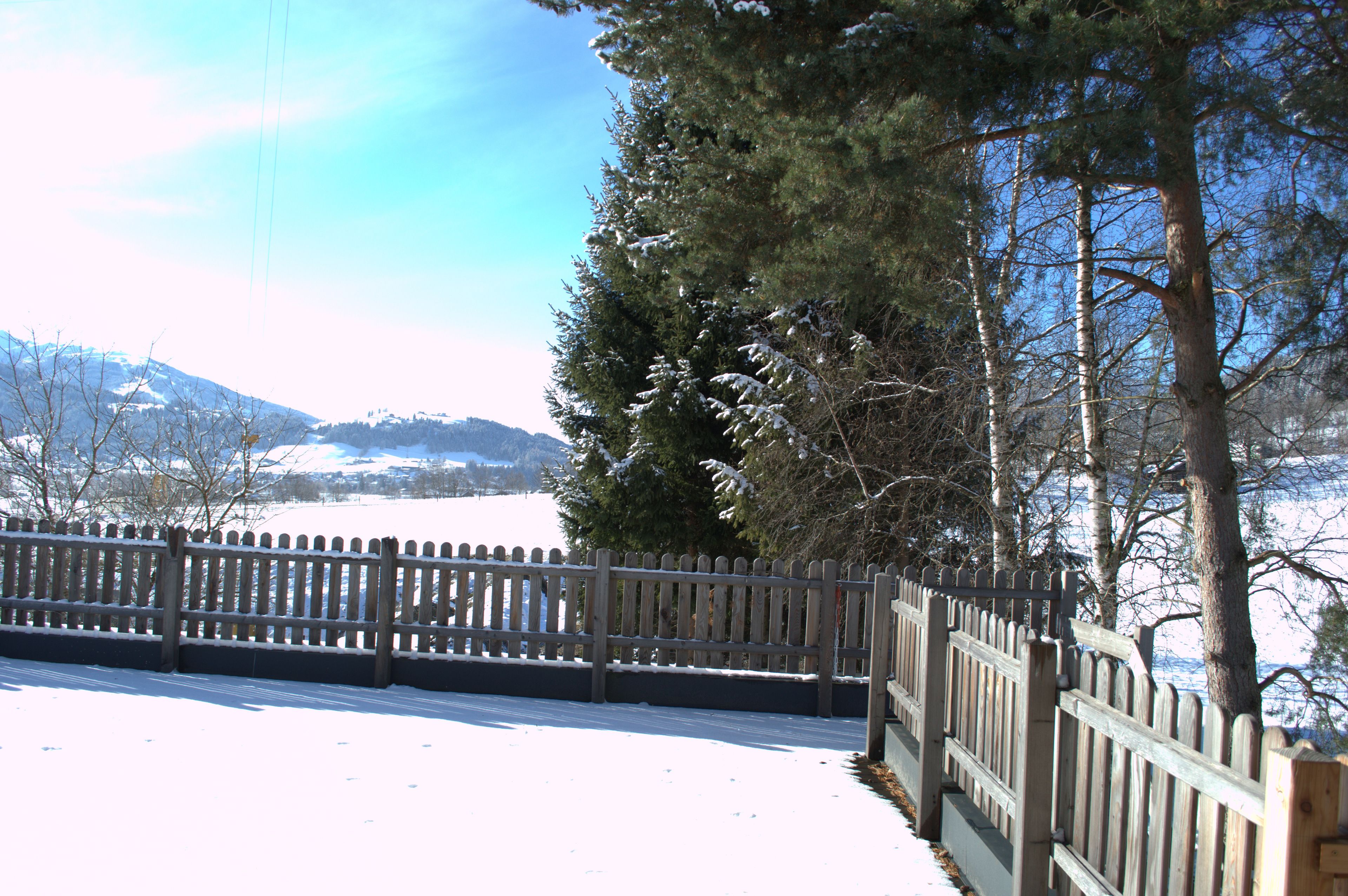 A snow-covered landscape with a wooden fence, trees, and a clear sky.