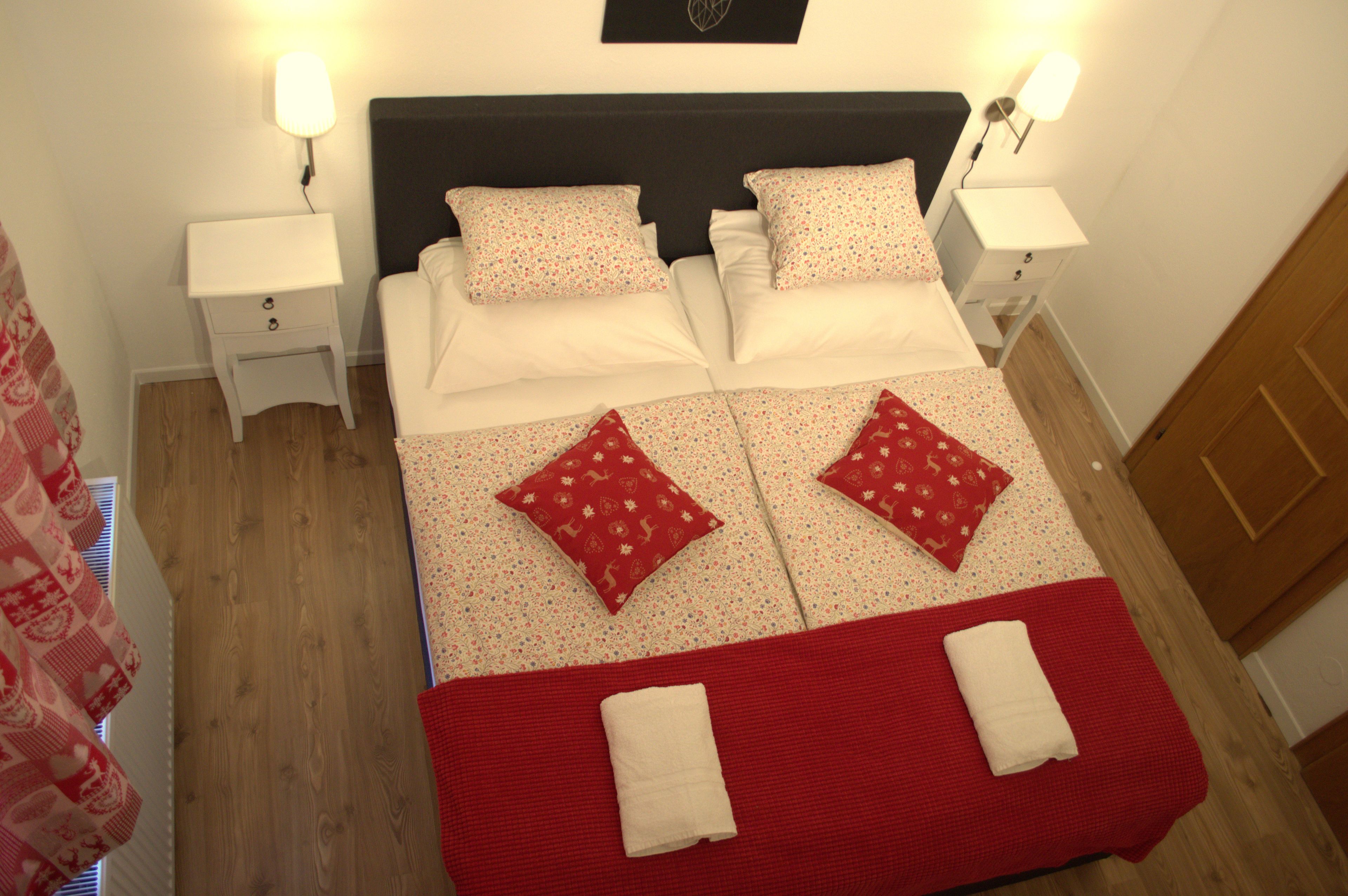 Una cama bien hecha con una colcha roja y blanca, dos almohadas y dos mesitas de noche con lámparas.