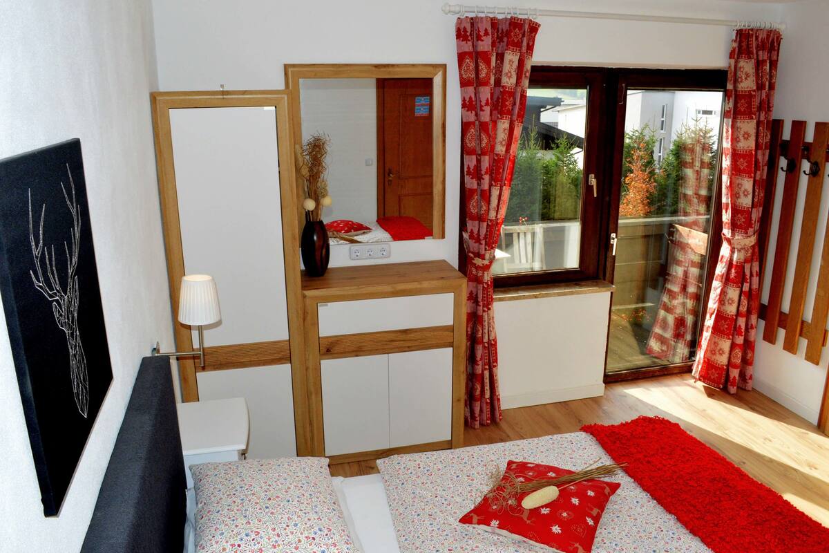 A bedroom with a bed, a bedside table, a mirror, a window with curtains, and a view of the outside.