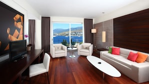 A modern hotel room with a large window offering a scenic view of the sea and mountains, a flat-screen TV, a sofa with colorful cushions, and a wooden floor. - No 81 Hotel (Bodrum)
