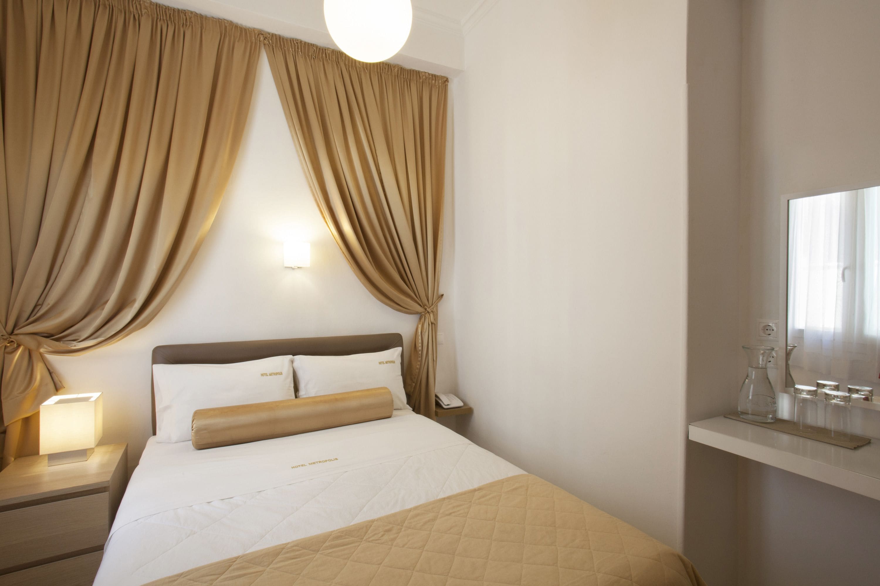 A hotel room with a bed, curtains, a nightstand with a lamp, a bedside table with a phone, and a mirror with a shelf above it.