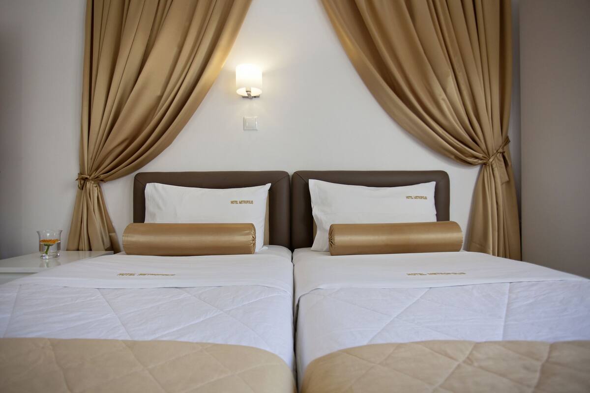 A hotel room with two beds, beige curtains, a wall-mounted light, and a small table with a glass and a flower.