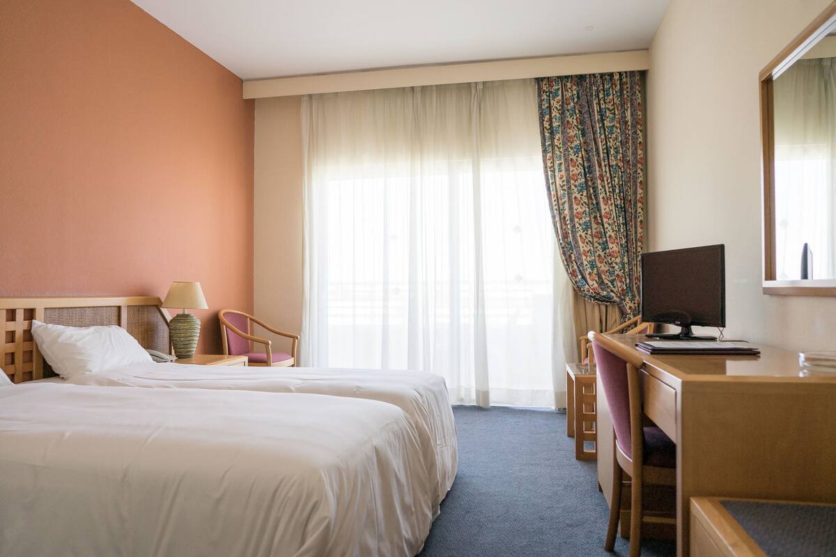 A hotel room with two beds, a desk, a chair, a lamp, and a television.