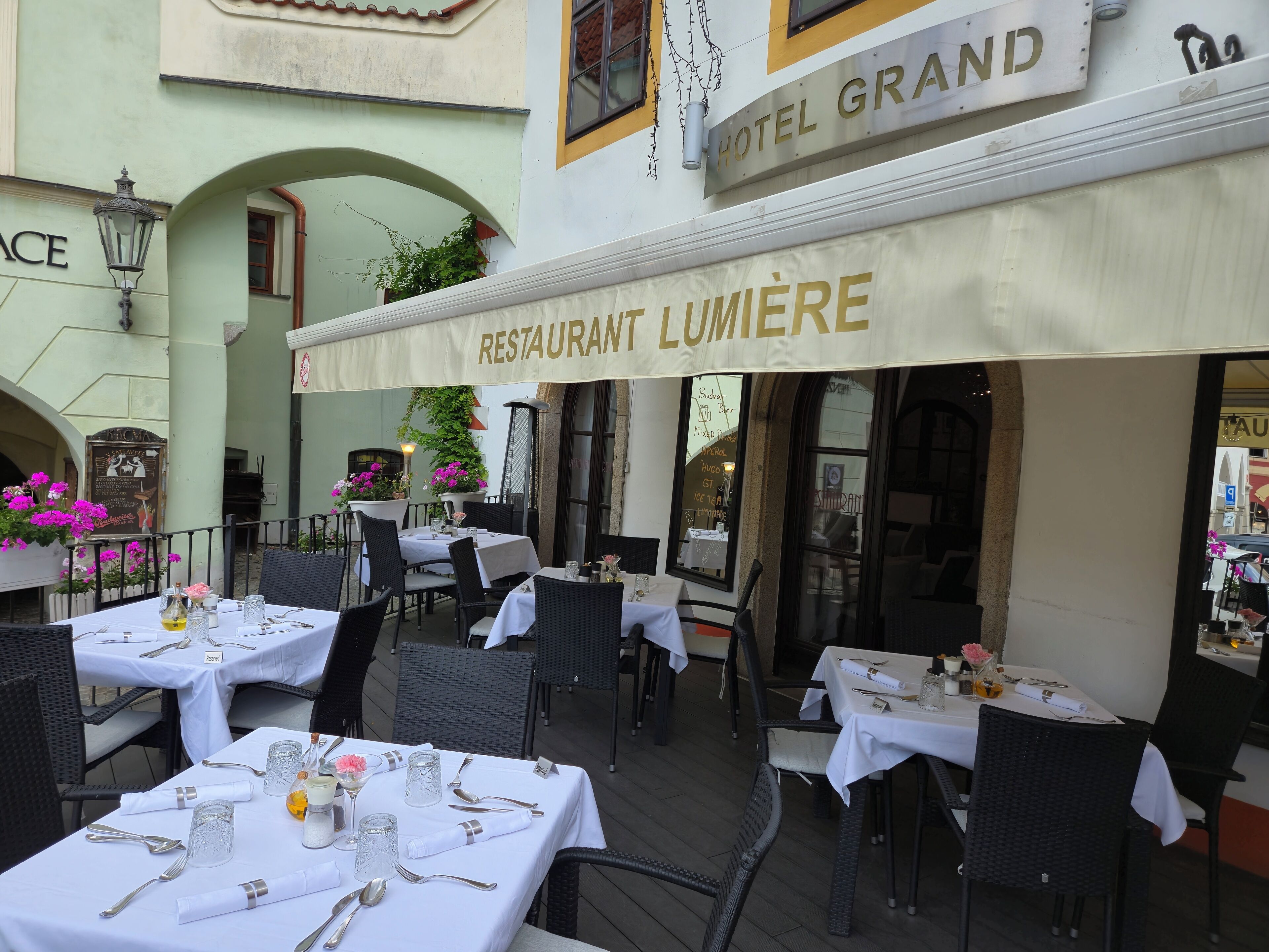 Un área de comedor al aire libre con mesas dispuestas para los huéspedes, un letrero que dice 'RESTAURANT LUMIÈRE' y el letrero 'HOTEL GRAND' en la parte superior.