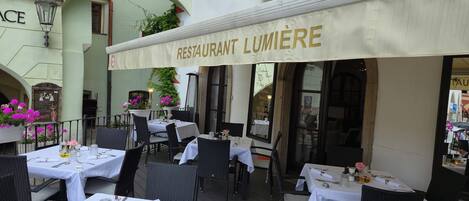 Un área de comedor al aire libre con mesas dispuestas para los huéspedes, un letrero que dice 'RESTAURANT LUMIÈRE' y el letrero 'HOTEL GRAND' en la parte superior.