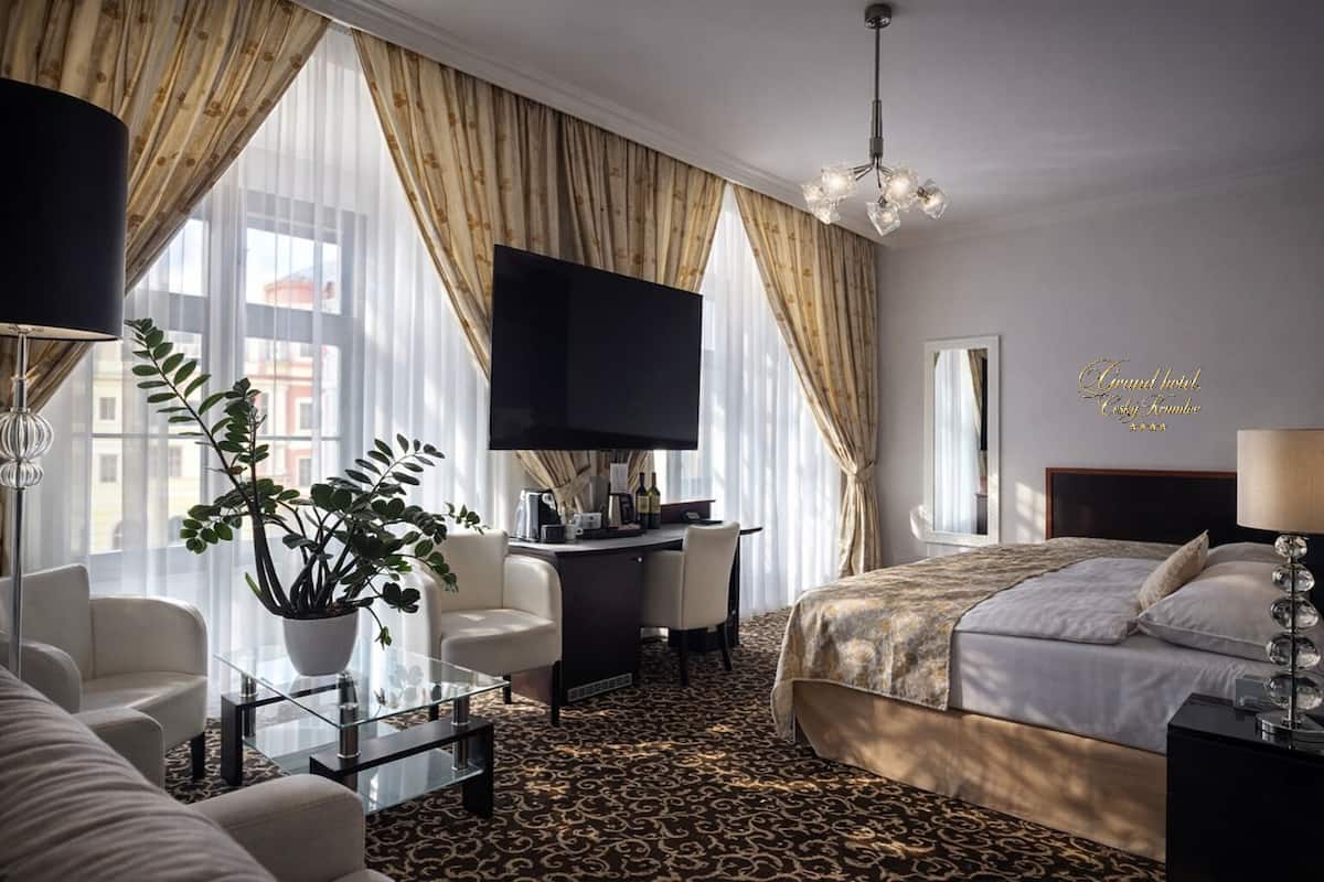 Habitación de hotel con cama, escritorio, televisor, sofá y un amplio ventanal con cortinas.
