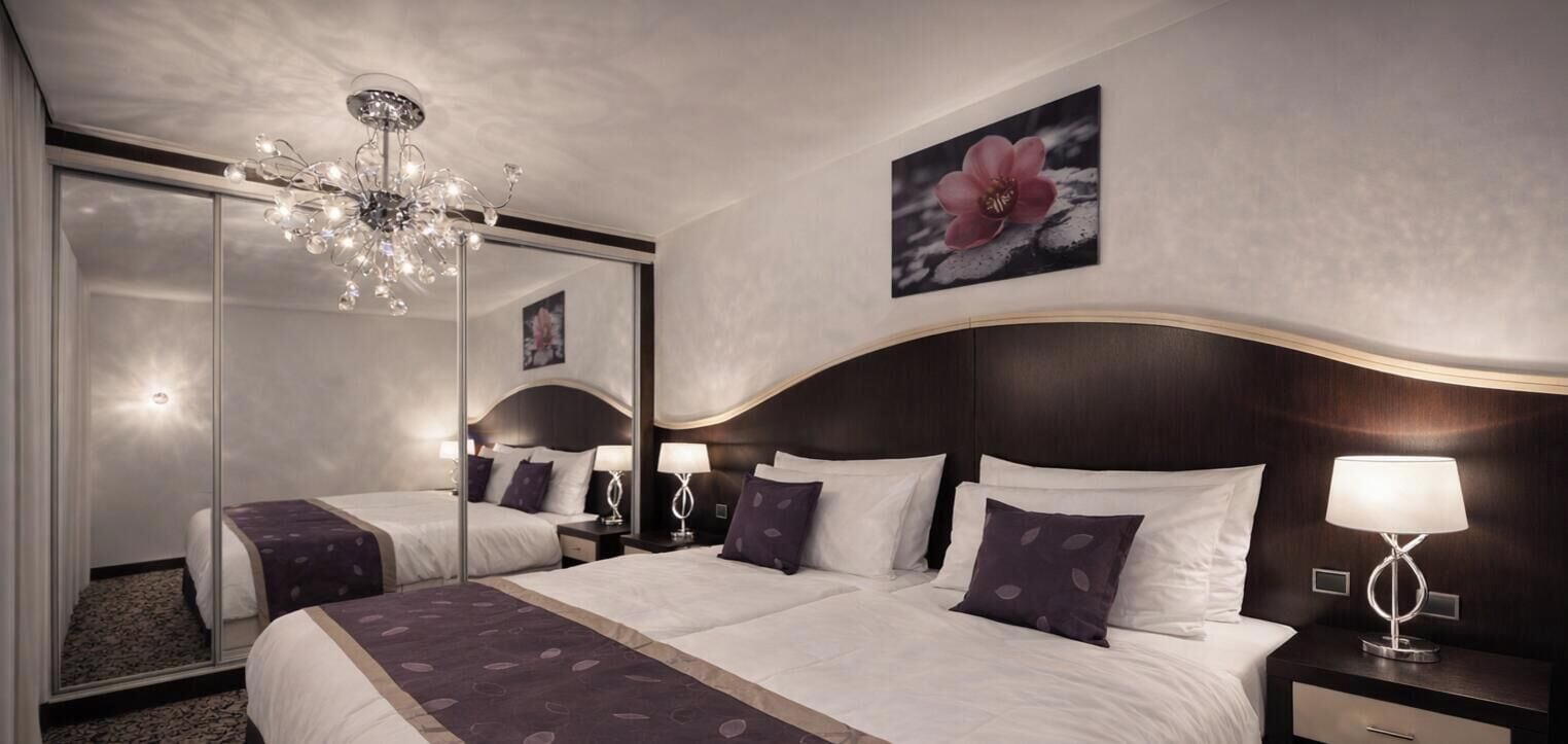 A hotel room with a large bed, a chandelier, a mirror, and a flower painting on the wall.