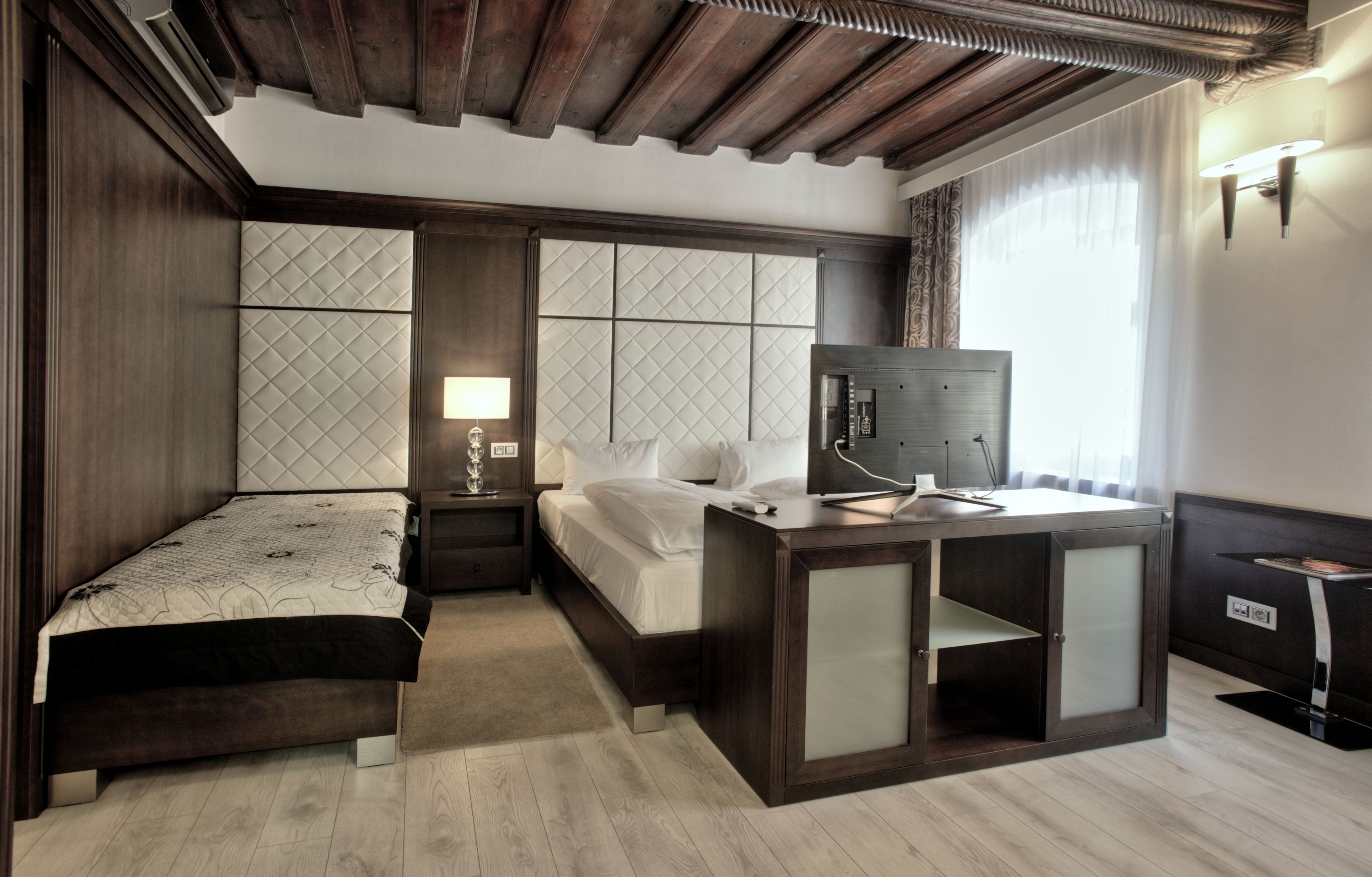 Una habitación de hotel con techo de madera, cama con cabecero acolchado, mesita de noche con lámpara, escritorio con espejo y armario.
