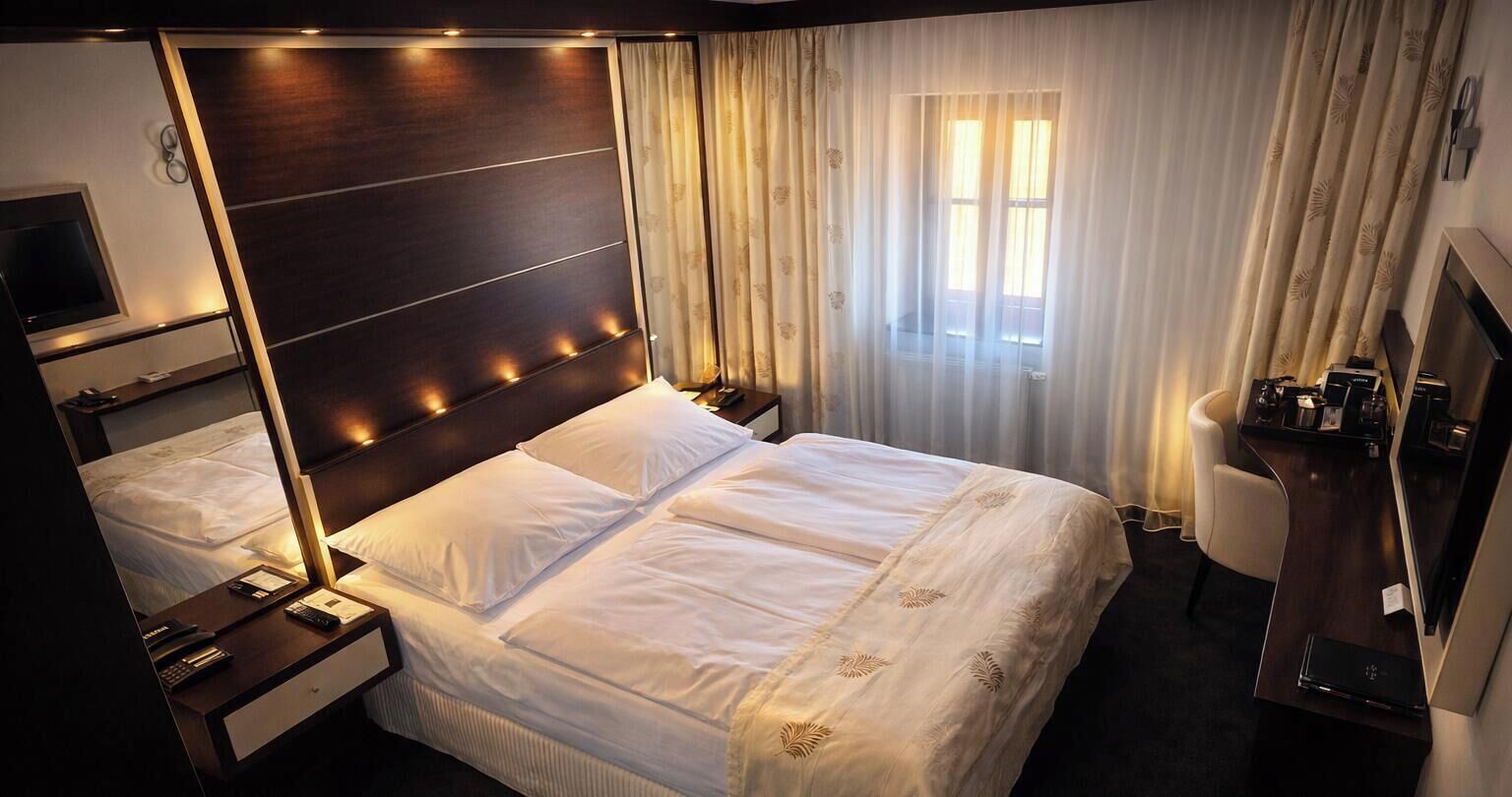 A hotel room with a large bed, bedside tables, a desk, a chair, a TV, and a window with curtains.