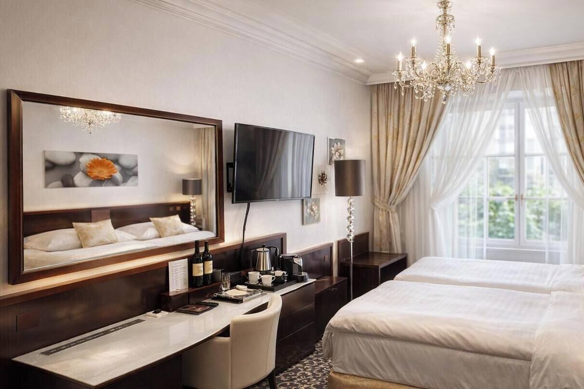 Una habitación de hotel con una cama grande, un escritorio con silla, televisor, candelabro y ventana con cortinas.