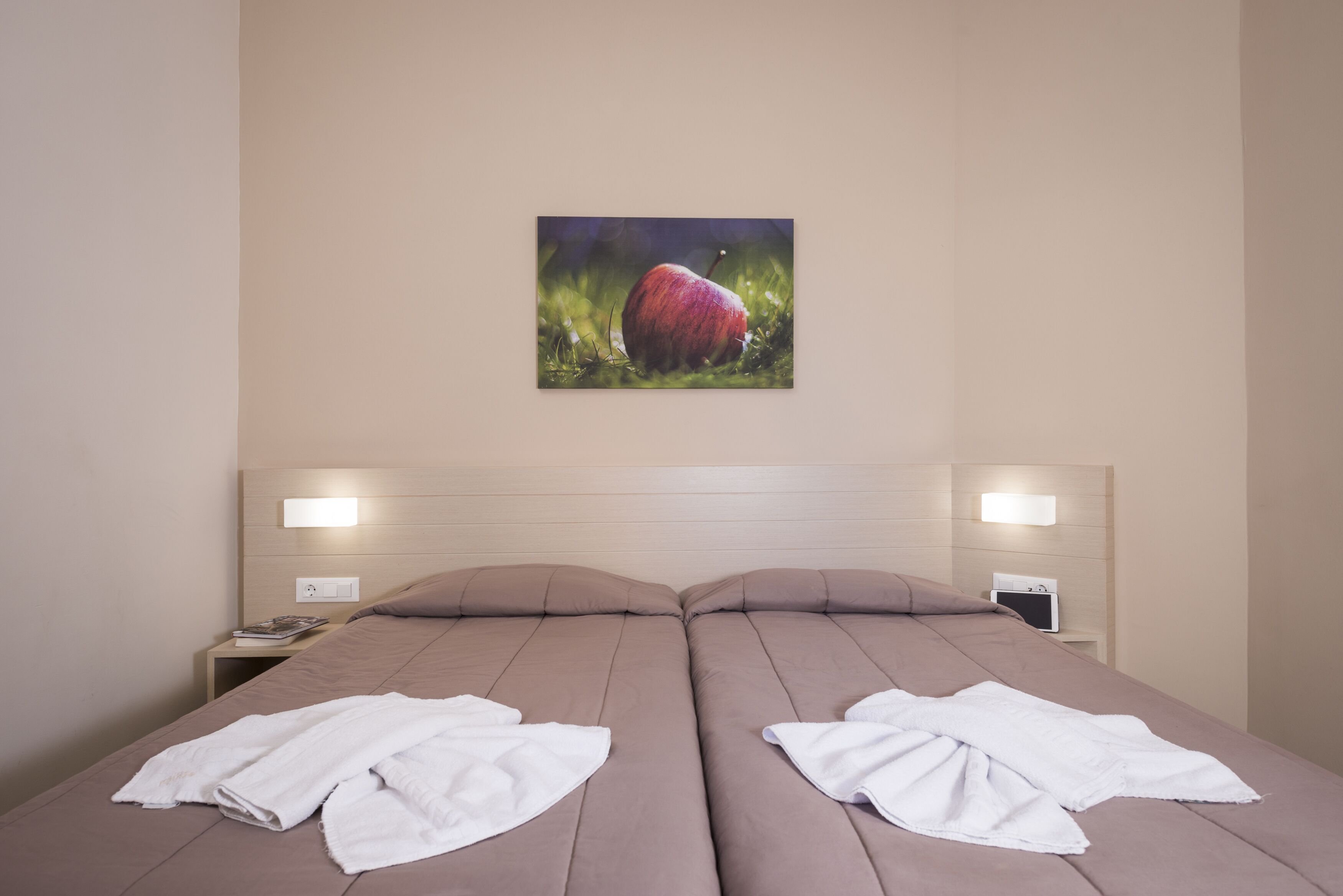 Un lit double avec deux serviettes blanches, un tableau représentant une pomme rouge accroché au mur, et des lampes de chevet.