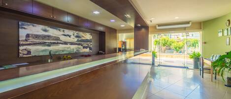 A spacious lobby with a long reception desk, a large wall-mounted artwork, and a glass door leading to an outdoor area with plants.