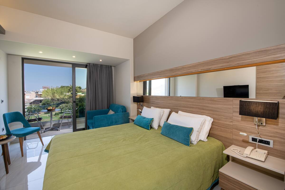 A modern hotel room with a large bed, a balcony with a table and chairs, a flat-screen TV, and a telephone.