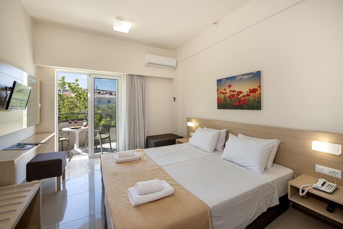 A hotel room with a bed, bedside table, and a desk with a chair. There is a window with a view of buildings and trees.
