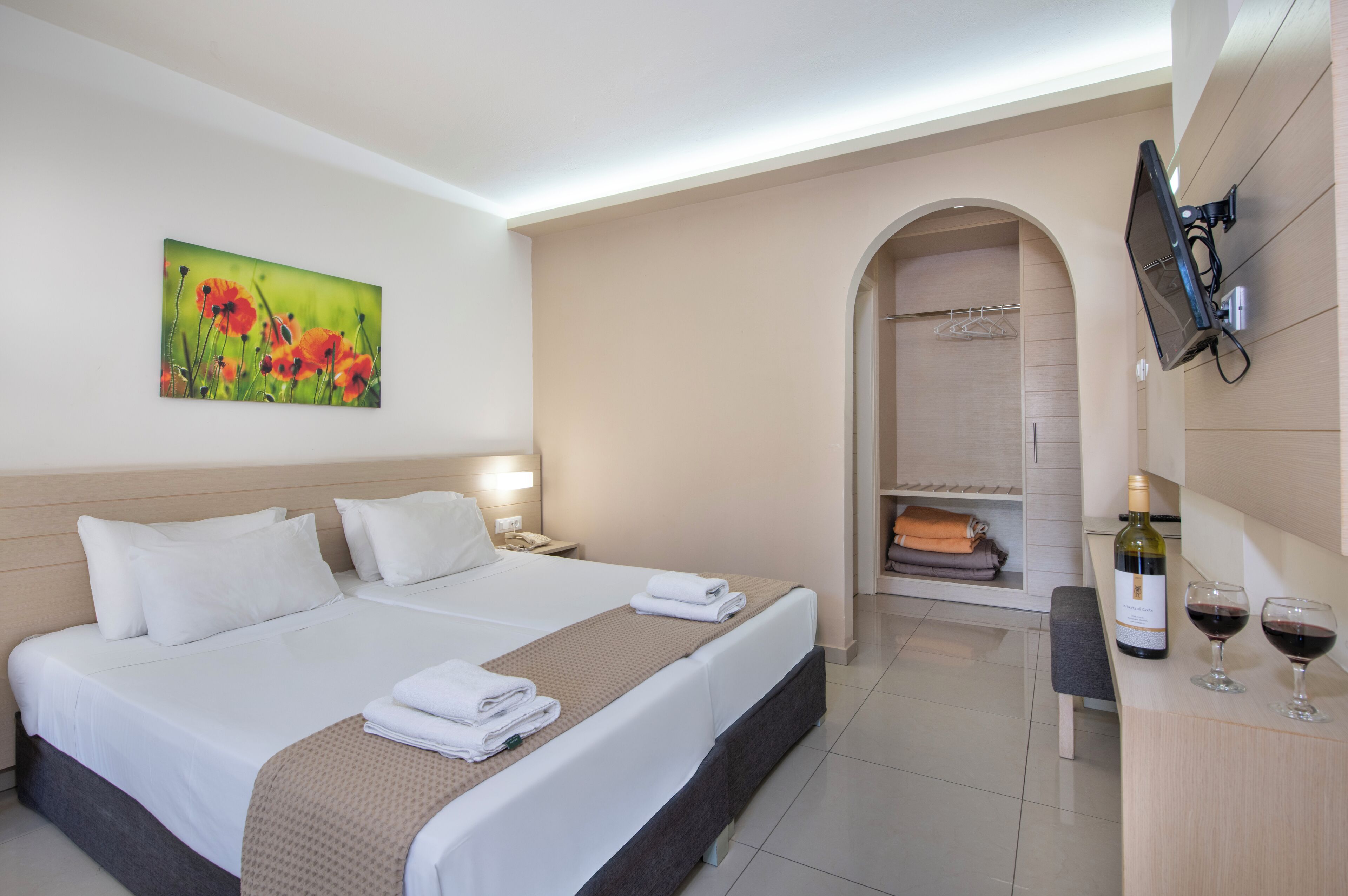 A hotel room with a large bed, a TV mounted on the wall, a wine bottle and glasses on a small table, and a painting of flowers on the wall.