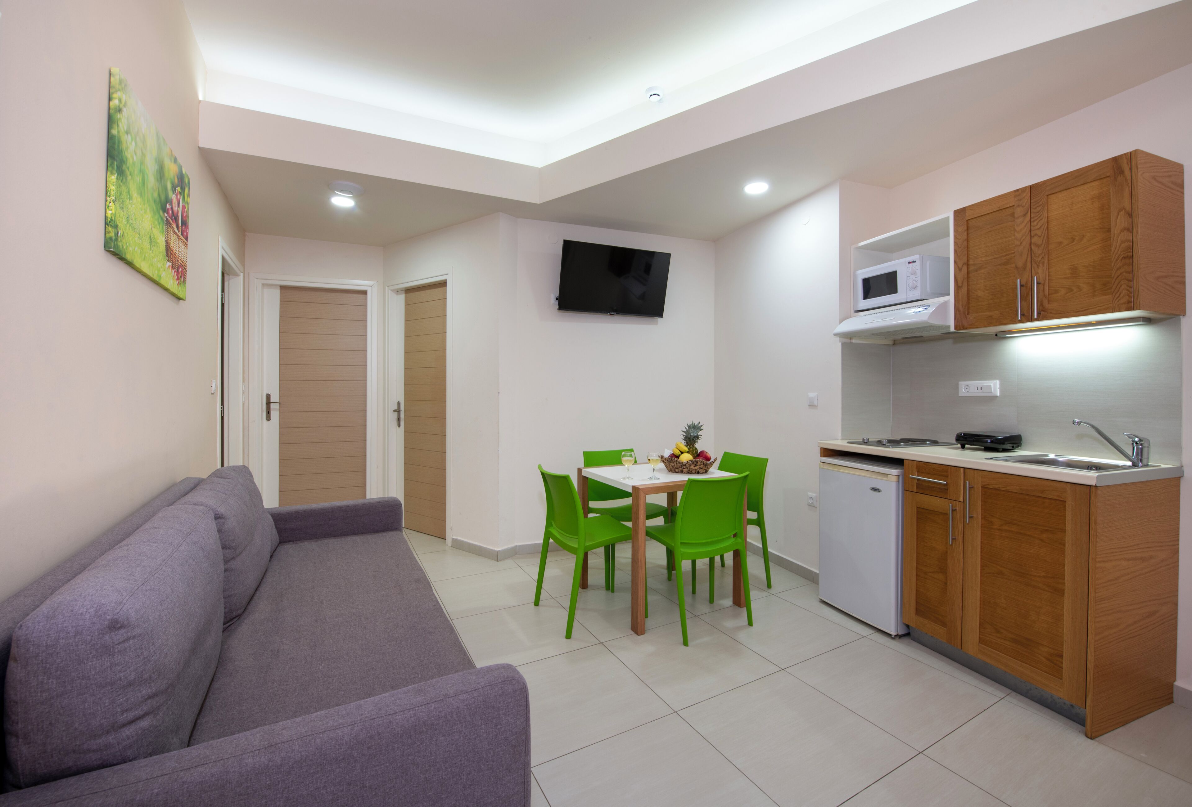 A compact living space with a sofa, a dining table with chairs, a TV mounted on the wall, and a kitchenette with cabinets and a microwave.