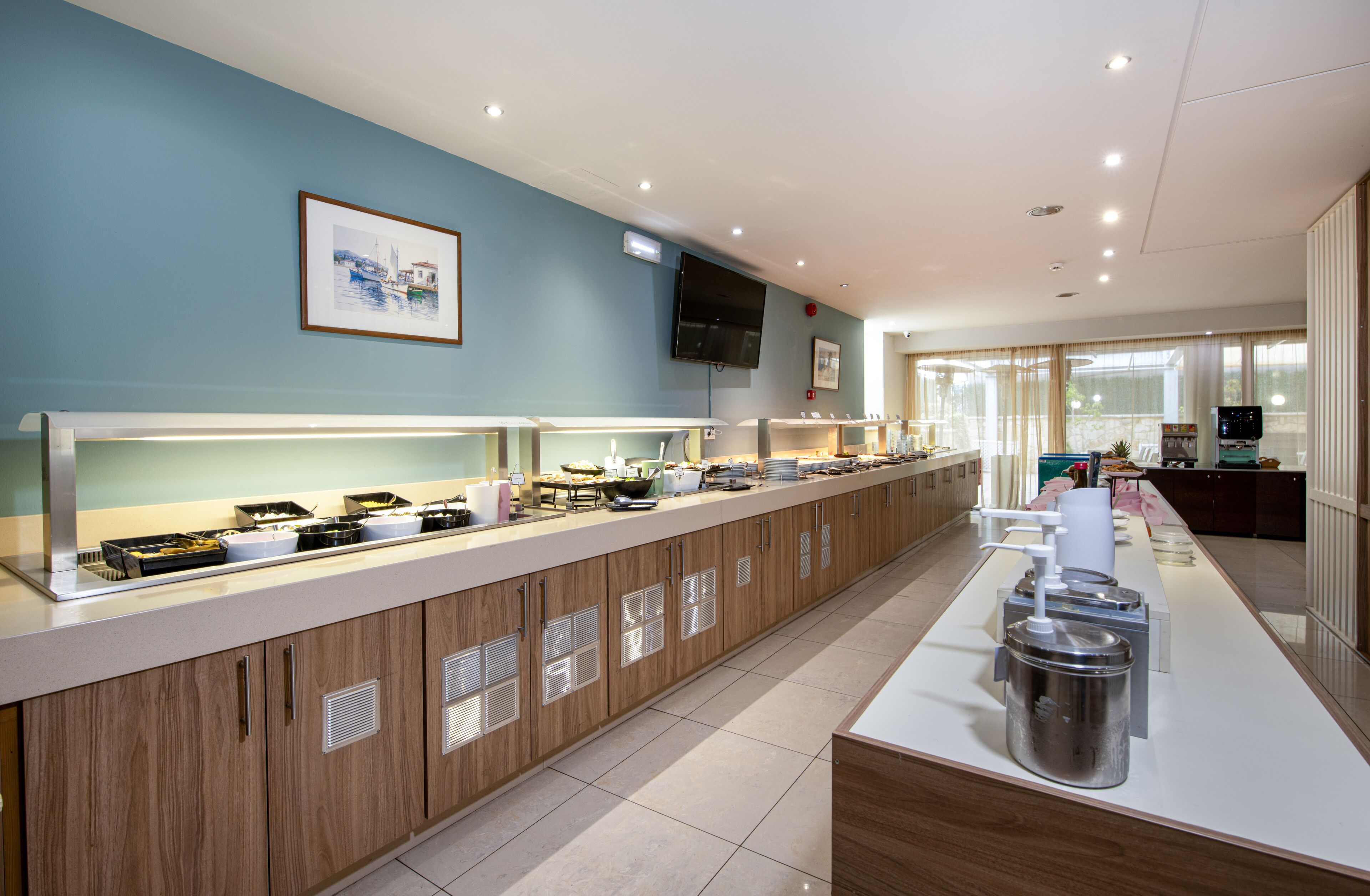 A modern hotel kitchen with a long counter, multiple food stations, and a television mounted on the wall.