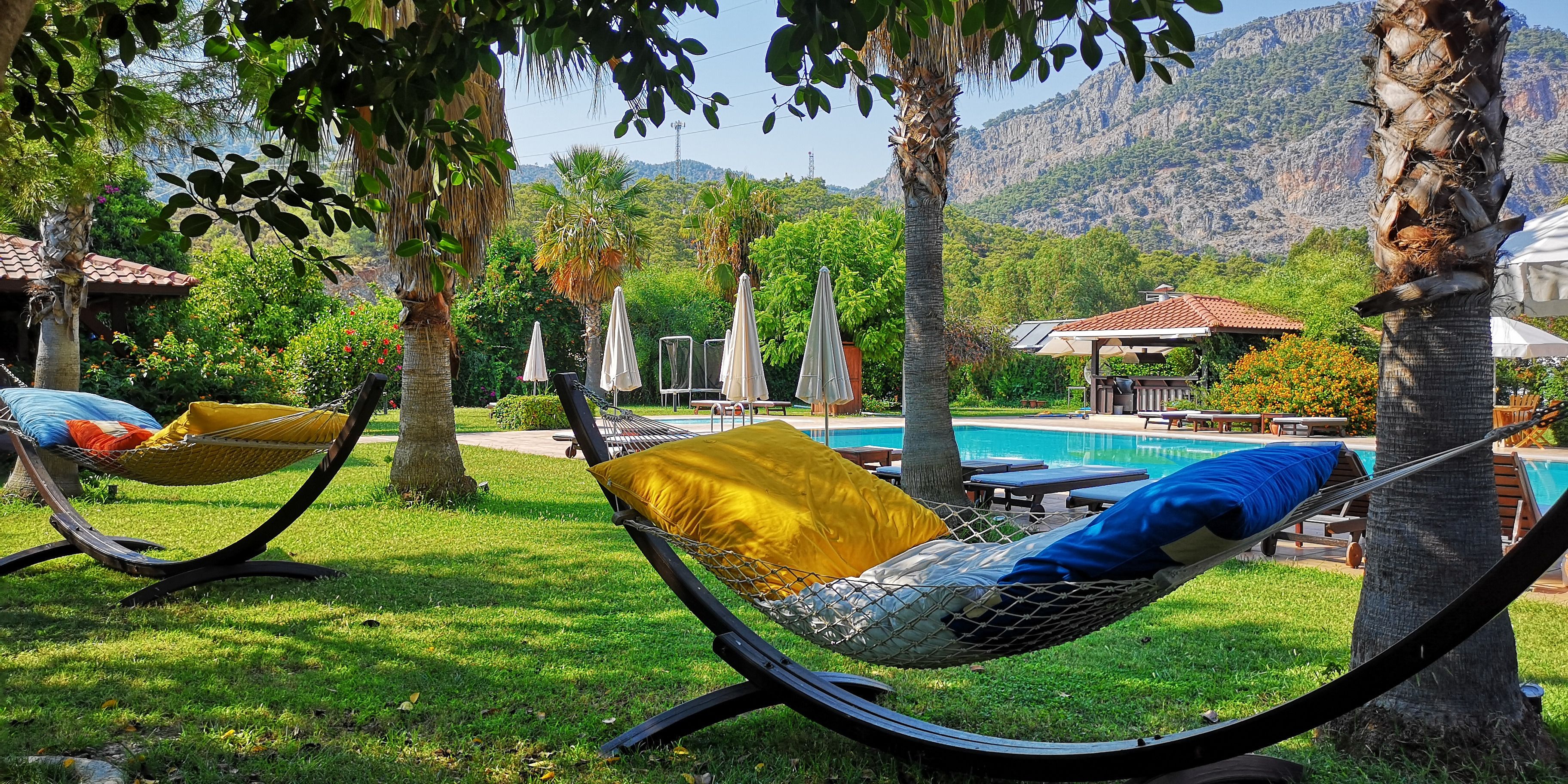 Un fauteuil suspendu avec un coussin jaune, un espace aménagé au bord de la piscine avec des chaises longues et des parasols, et un paysage montagneux en arrière-plan.