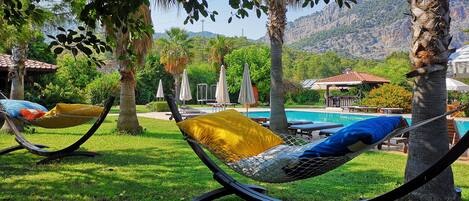 Un fauteuil suspendu avec un coussin jaune, un espace aménagé au bord de la piscine avec des chaises longues et des parasols, et un paysage montagneux en arrière-plan.