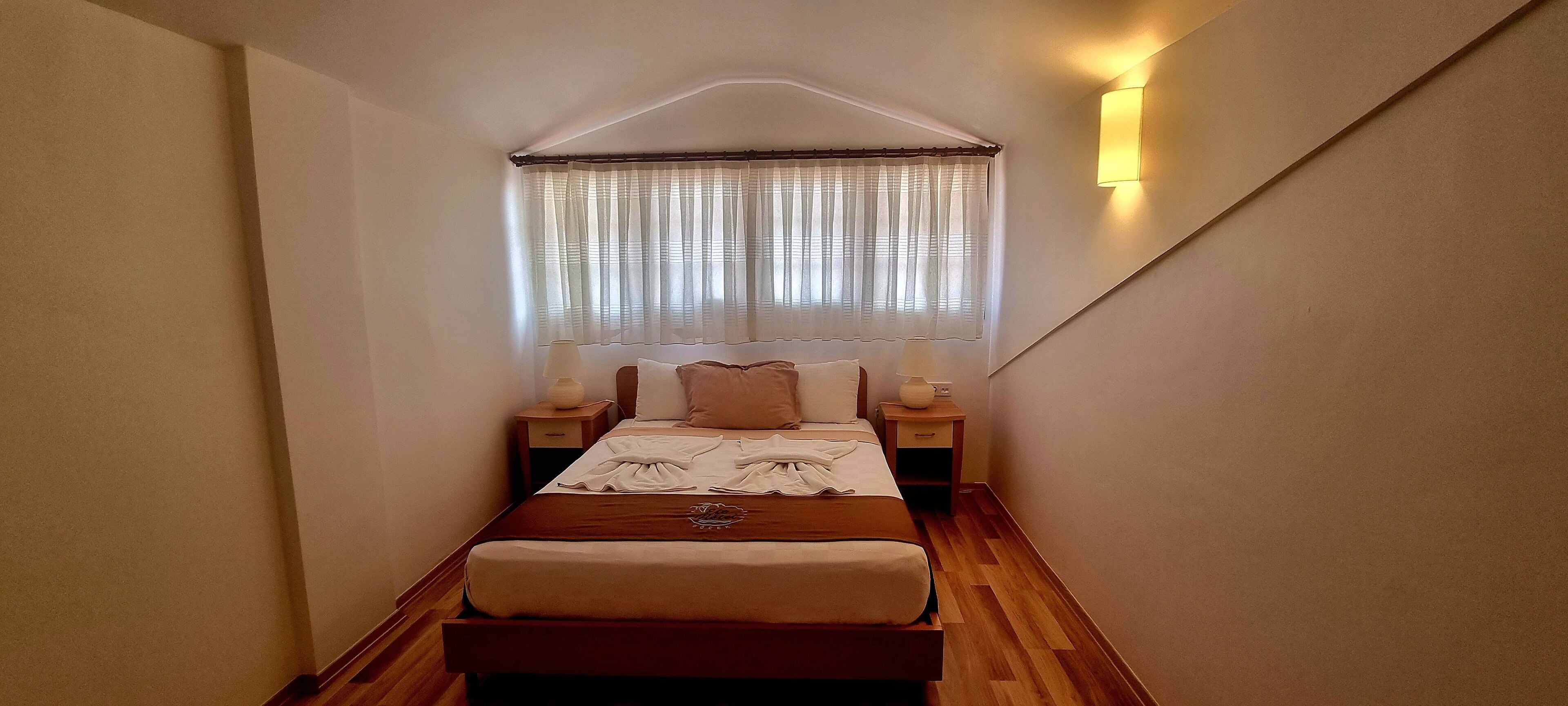 A narrow, well-lit bedroom with a wooden floor, a bed with a headboard, two bedside tables with lamps, and a window with sheer curtains.