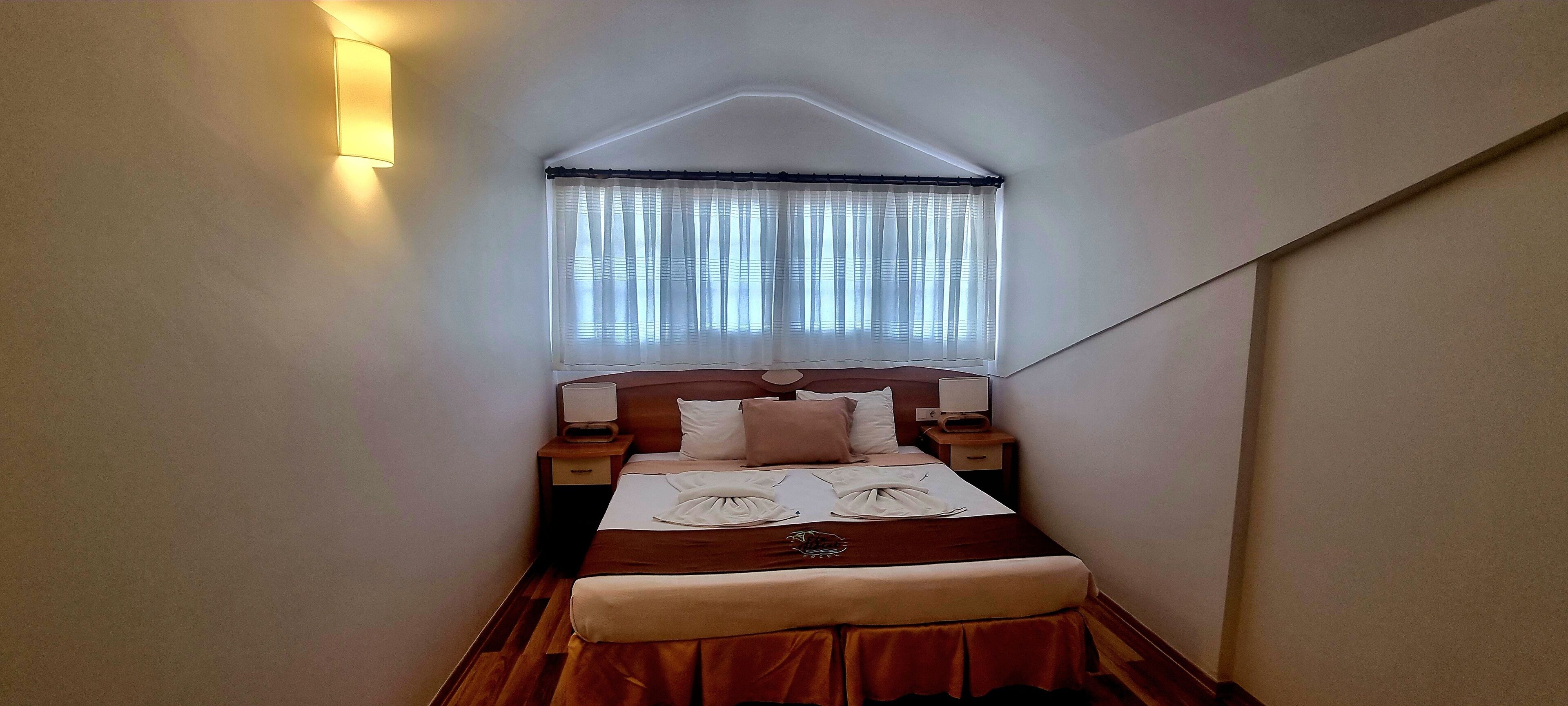 Una habitación estrecha y con techo inclinado, con cama, mesitas de noche y una ventana con cortinas.