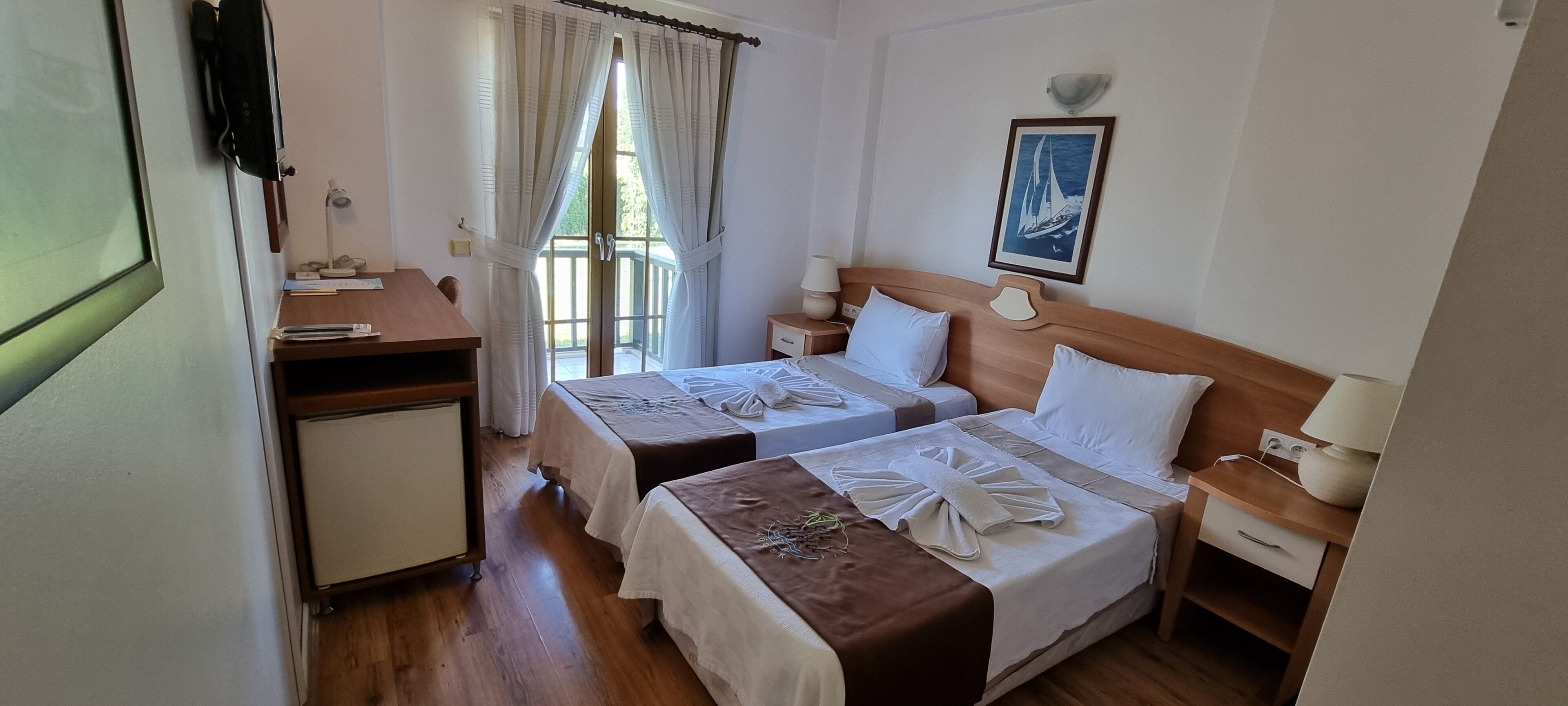 A hotel room with two beds, a desk, a small fridge, and a window with curtains.