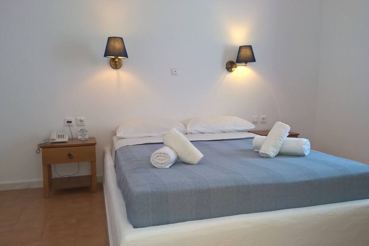 Un lit bien fait, avec du linge de lit blanc et des serviettes pliées, deux lampes fixées au mur, une table de chevet avec un téléphone et un panneau, et une table de nuit en bois.
